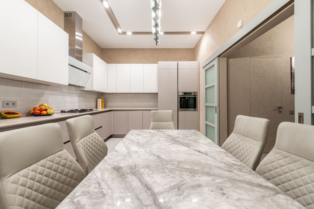 Contemporary kitchen in beige and white colors with minimalist cabinets and marble table with soft chairs under modern lamp